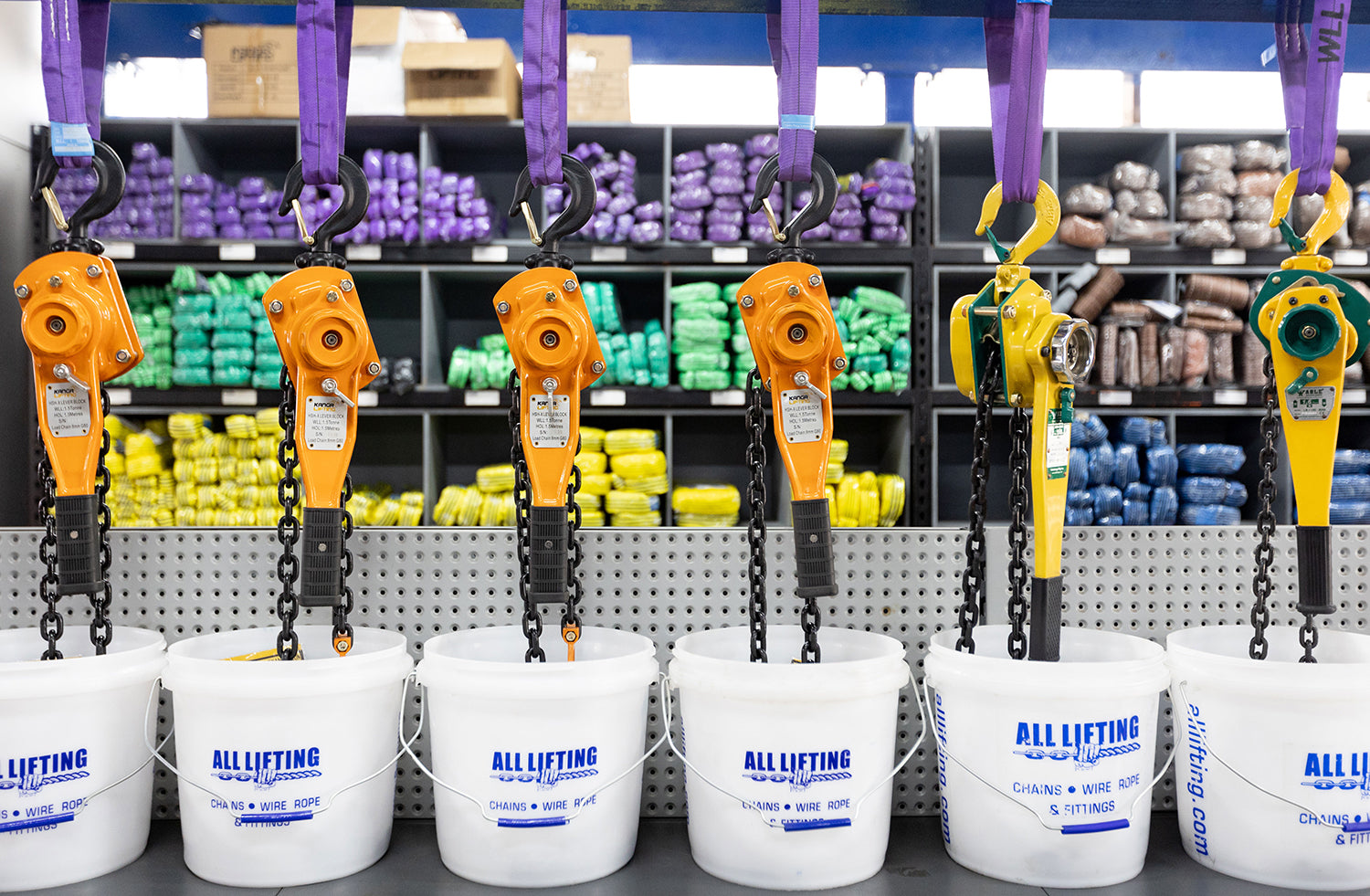 A row of orange and yellow chain hoists hanging from purple straps above white buckets printed with the brand name 'ALL LIFTING', set against a warehouse background with shelves of packaged goods.