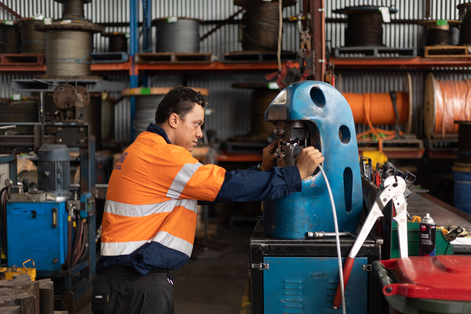 A worker in an orange high-visibility safety jacket with reflective stripes operates a blue industrial machine pressing a wire rope sling inside a workshop or factory